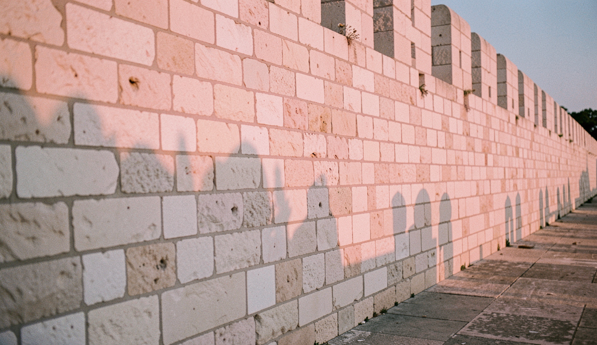 Longue muraille de pierre claire éclairée de côté en fin de journée, chaque bloc taillé projette une ombre courte et la couleur vire au rose