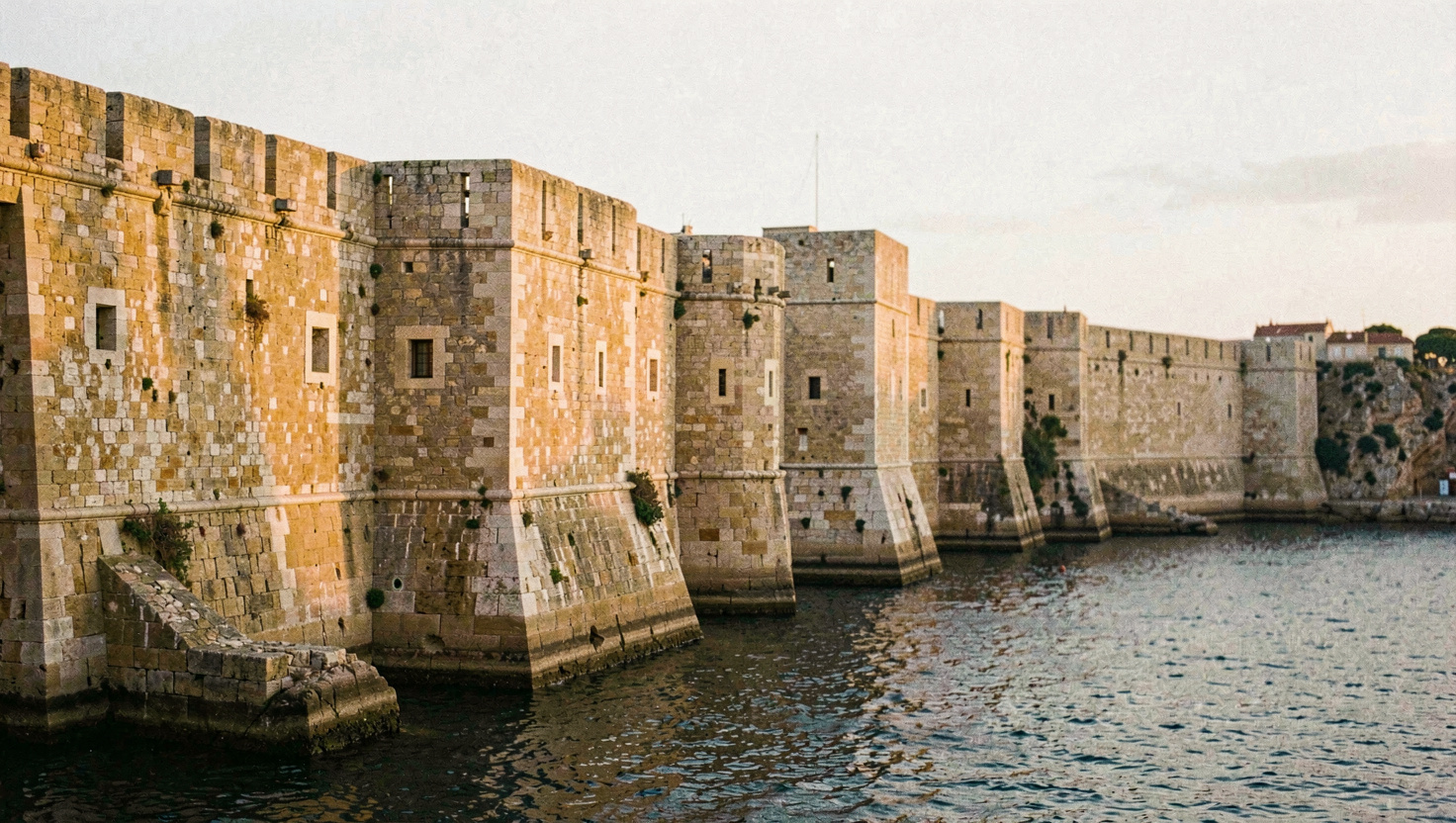 Bastions de pierre dorée surplombant un port naturel, la lumière rasante du soir accentue les assises taillées et les meurtrières